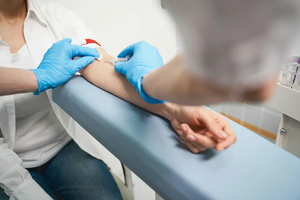 A healthcare worker wearing blue gloves draws blood from a person's arm using a syringe. The person is seated with their arm resting on a padded surface.