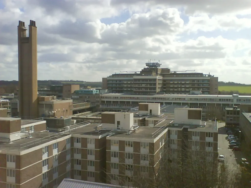 A large university campus with several multi storey, rectangular buildings and a tall, narrow clock tower, under a partly cloudy sky with green fields in the background.